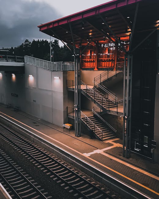 A modern East Croydon station platform during dusk, showing an external staircase with metal handrails and concrete steps leading to an upper level with a glass-fronted structure illuminated by warm orange lighting. The platform features tactile yellow safety lines along the edge and is adjacent to railway tracks with visible metal rails and sleepers. The station's exterior wall is constructed of smooth grey concrete with integrated lighting fixtures, while the surrounding environment includes trees and cloudy sky. The scene reflects aspects of urban transport infrastructure relevant to house removals and furniture transport logistics, as arranged by Removal Van Croydon, especially when managing access challenges during home relocation or packing and moving processes.