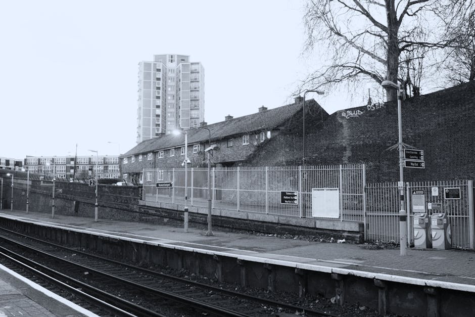 A modern East Croydon station platform during dusk, showing an external staircase with metal handrails and concrete steps leading to an upper level with a glass-fronted structure illuminated by warm orange lighting. The platform features tactile yellow safety lines along the edge and is adjacent to railway tracks with visible metal rails and sleepers. The station's exterior wall is constructed of smooth grey concrete with integrated lighting fixtures, while the surrounding environment includes trees and cloudy sky. The scene reflects aspects of urban transport infrastructure relevant to house removals and furniture transport logistics, as arranged by Removal Van Croydon, especially when managing access challenges during home relocation or packing and moving processes.